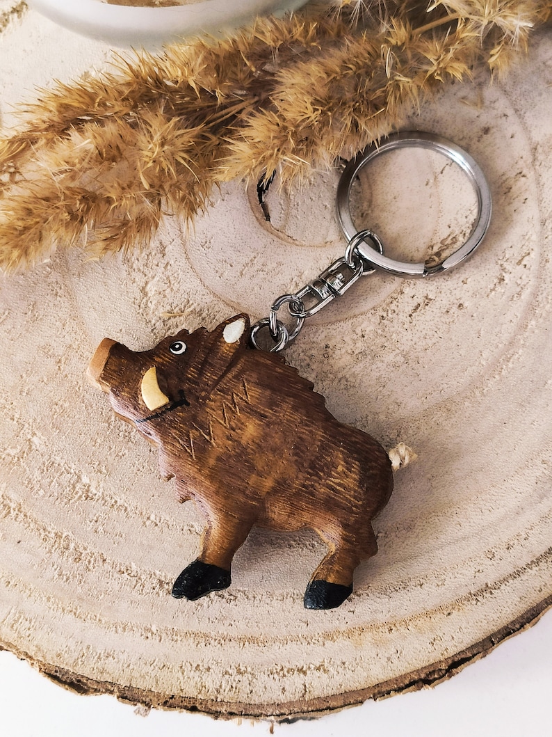 May include: Wooden boar keychain with a brown and black painted finish. The keychain has a silver metal ring and chain.