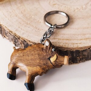 May include: A wooden boar keychain with a painted face and a metal key ring. The boar is brown with black hooves and a yellow tusk.