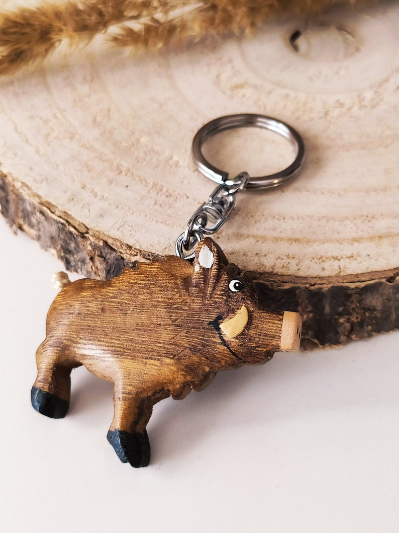 May include: A wooden boar keychain with a painted face and a metal key ring. The boar is brown with black hooves and a yellow tusk.