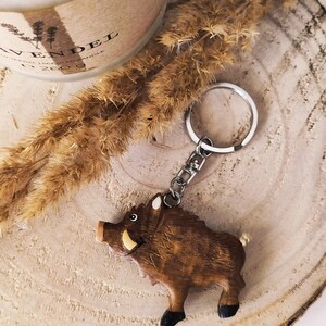 May include: Wooden wild boar keychain with a brown and black finish. The keychain is attached to a silver key ring.