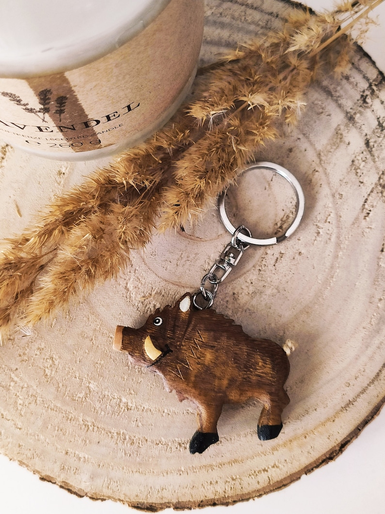 May include: Wooden wild boar keychain with a brown and black finish. The keychain is attached to a silver key ring.
