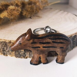 May include: A wooden boar keychain with black stripes, attached to a silver key ring. The boar is standing on a wooden surface.