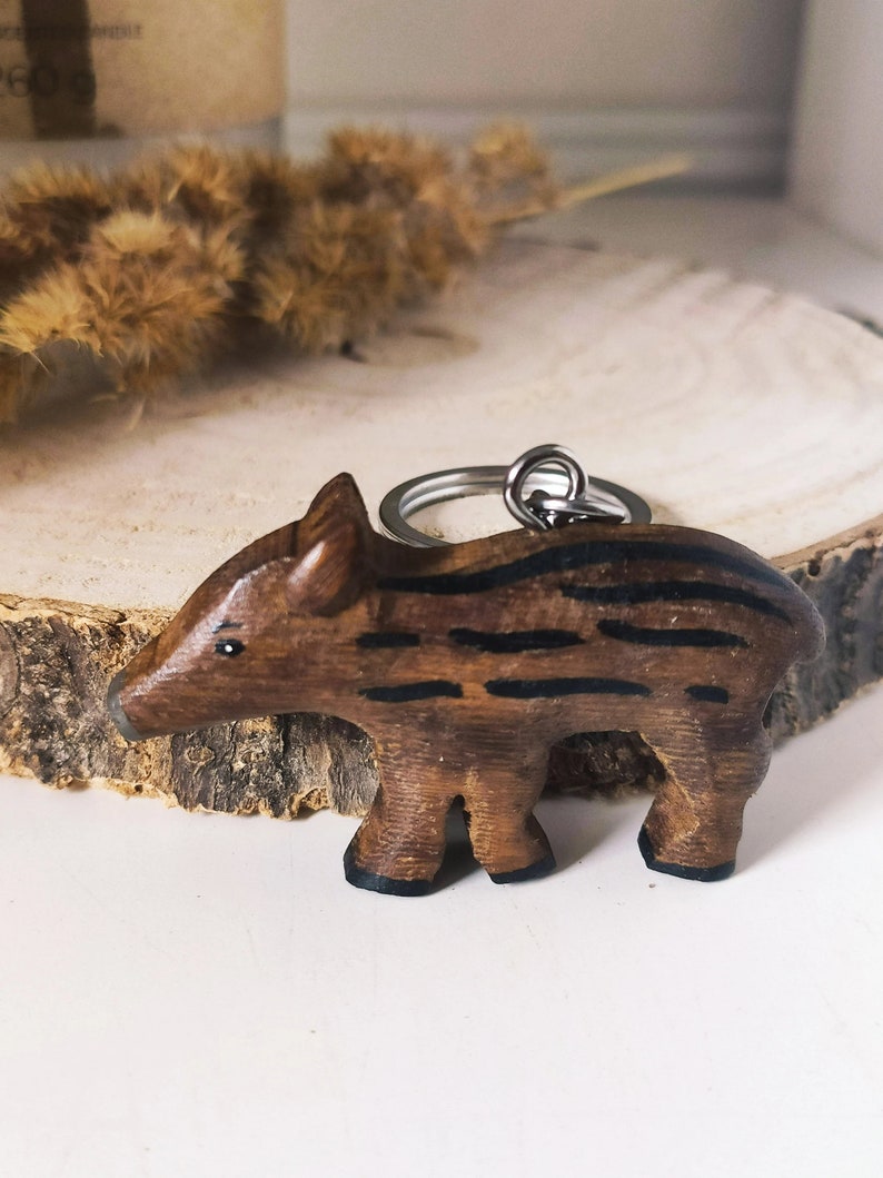 May include: A wooden boar keychain with black stripes, attached to a silver key ring. The boar is standing on a wooden surface.