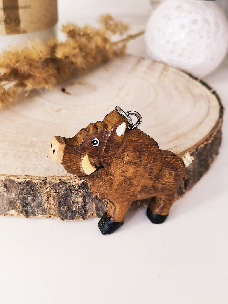 May include: A wooden wild boar keychain with a brown body, black hooves, and a white tusk. The keychain has a silver loop for attaching to a bag or keys.