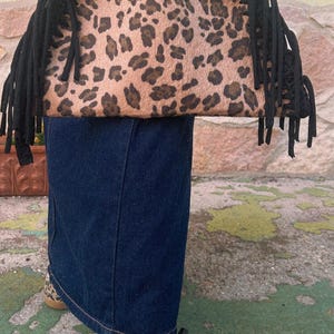 Può includere: Una borsa con motivo leopardato beige e marrone, dotata di un manico superiore all'uncinetto nero e lunghe frange nere sui lati. La borsa è abbinata a scarpe da ginnastica basse con una stampa leopardata coordinata e inserti laterali bianchi.