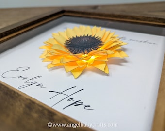 May include: A framed paper craft of a yellow sunflower with a black center. The frame has a white background with the text "Evelyn Hope" and "Be sunshine" written in black cursive. The bottom of the frame has the website "www.angelbabycrafts.com" printed in black.