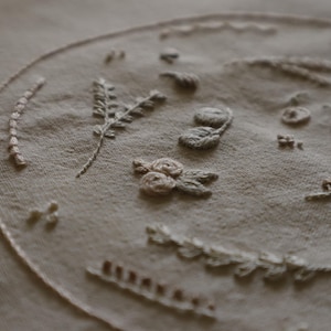Puede incluir: Primer plano de una tela beige con bordados detallados. El bordado presenta varios diseños florales y botánicos, incluyendo trigo, flores y patrones circulares. Las puntadas son de un color beige similar, creando un efecto sutil y texturizado.