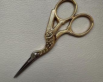 May include: Gold-colored scissors with a bird design. The handles are decorated with intricate details. The blades are sharp and pointed.