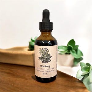 May include: A brown glass bottle of Comfrey Herbal Tincture with a black dropper. The label features a botanical illustration and the text "Comfrey Herbal Tincture Symphytum x Uplandicum 2 oz."