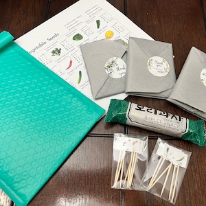 May include: An assortment of items, including seed packets, a teal bubble mailer, and a snack bar, are arranged on a wooden surface. The seed packets are wrapped in gray paper and sealed with floral "thank you" stickers. A printed sheet lists vegetable seeds.