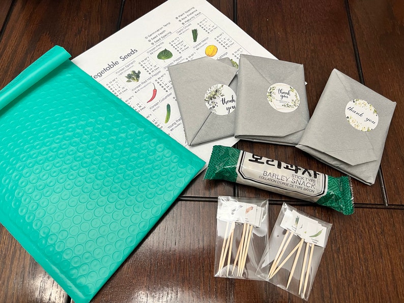 May include: An assortment of items, including seed packets, a teal bubble mailer, and a snack bar, are arranged on a wooden surface. The seed packets are wrapped in gray paper and sealed with floral "thank you" stickers. A printed sheet lists vegetable seeds.