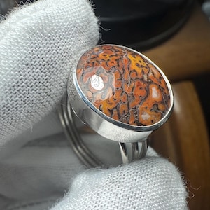 Op de afbeelding: Een zilveren ring met een grote, ovaalvormige cabochonsteen met een complex, webachtig patroon in tinten oranje, geel en donkerbruin. De steen is gezet in een gladde, gepolijste zilveren zetting en de ringband bestaat uit drie dunne zilveren draden.