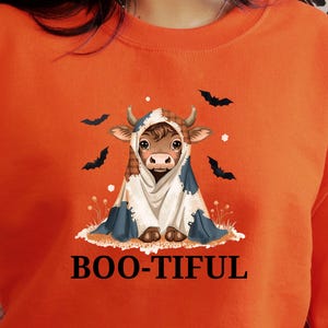 Op de afbeelding: Oranje sweatshirt met een Halloween-thema. De afbeelding toont een cartoon koe in een kostuum, met een cape en een capuchon. De tekst "BOO-TIFUL" staat eronder gedrukt. Zwarte vleermuis silhouetten zijn ook aanwezig.