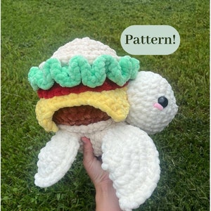 May include: A white crocheted turtle with a hamburger on its back. The hamburger has green lettuce, red tomato, yellow cheese, and a brown patty. The turtle has black eyes and a pink nose.