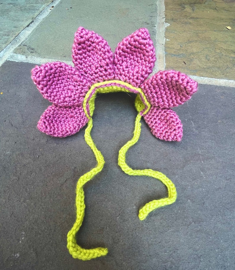Pode incluir: Um chap&eacute;u de croch&ecirc; rosa em forma de flor com la&ccedil;os de croch&ecirc; verde. O chap&eacute;u tem a forma de uma flor com cinco p&eacute;talas.