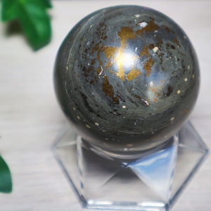 Könnte beinhalten: Eine dunkelgraue Kugel mit Goldflocken, die auf einem klaren, geometrischen Ständer ruht. Die Kugel hat ein marmoriertes Aussehen mit Andeutungen von Blau und Weiß. Der Hintergrund umfasst grünes Laub.