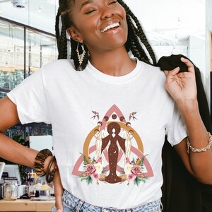 May include: A white t-shirt with a pink, gold, and brown graphic design featuring a woman with a halo and a floral pattern. The design is reminiscent of a Celtic knot.