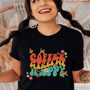May include: A black t-shirt featuring a retro-style graphic that reads "Coffee Makes Me Happy" in colourful letters. The graphic also includes flowers, butterflies, and a coffee cup.