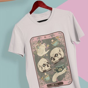 May include: A white t-shirt with a graphic of a tarot card featuring three skulls, a teapot, and teacups. The card is titled "The Tea" and has the number "II." on it.