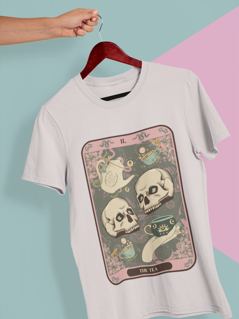 May include: A white t-shirt with a graphic of a tarot card featuring three skulls, a teapot, and teacups. The card is titled "The Tea" and has the number "II." on it.