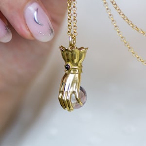 May include: A gold necklace with a pendant shaped like a hand holding a clear crystal orb. The hand is adorned with a small red gemstone on the wrist.