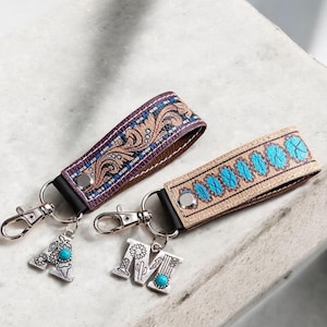 May include: Two keychains with decorative straps and silver-tone hardware. One strap is purple with a paisley design, the other is tan with blue accents. Each keychain has a letter charm with turquoise details.