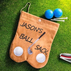 May include: A tan golf ball sack with the text "JASON'S BALL SACK" and a golf club illustration. Two golf balls and tees are next to the sack on a green surface. The sack has two golf ball patches.