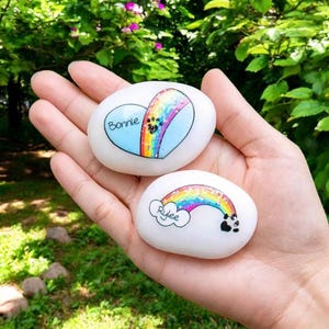 May include: Two white painted stones with rainbow and heart designs. One stone has a blue heart with the name "Bonnie" and a rainbow. The other stone has a rainbow with the name "Rylee" and a cloud.