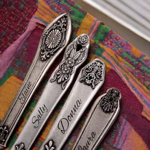 May include: Four silver spoons with ornate handles and engraved names: Elaine, Sally, Donna, and Laura. The spoons are arranged on a colorful patterned fabric.