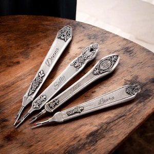 May include: Four silver seam rippers with ornate floral designs and personalized names: Donna, Sally, Elaine, and Laura. Each tool has a pointed tip with a red bead. The rippers are arranged on a wooden surface.