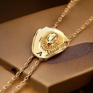 May include: A gold-colored bolo tie featuring a cowboy hat pendant. The hat has intricate detailing and the letter "A" is visible on the hat. The tie has a chain design.