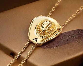 May include: A gold-colored bolo tie featuring a cowboy hat pendant. The hat has intricate detailing and the letter "A" is visible on the hat. The tie has a chain design.