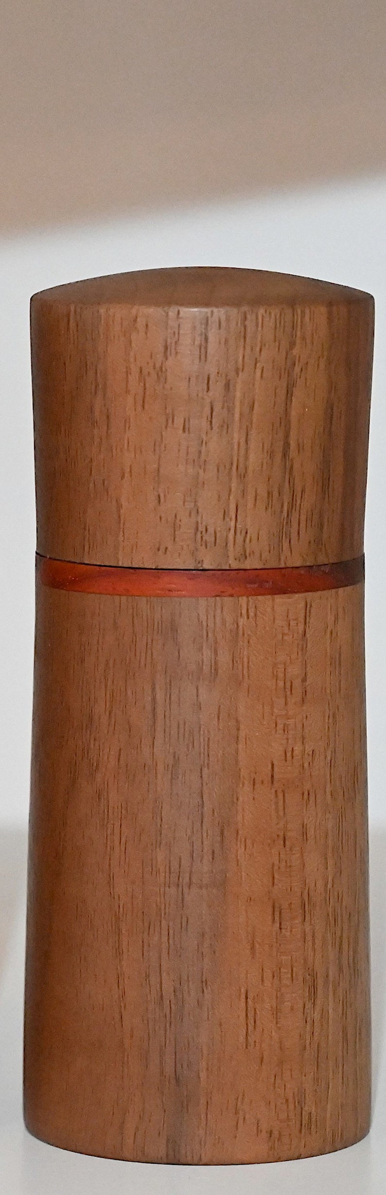 May include: A cylindrical wooden container with a red band around the middle. The container is made of a light brown wood with a natural grain pattern.