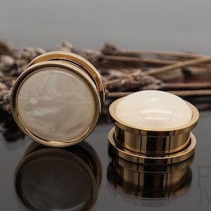 Peut inclure: Une paire d'écarteurs d'oreilles dorés avec des inserts blancs marbrés. Les écarteurs sont en métal et ont une finition lisse et polie.