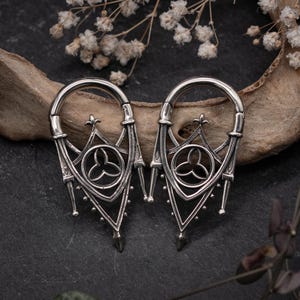 Puede incluir: Un par de pendientes de aro de plata de estilo gótico con detalles intrincados. Los pendientes presentan un diseño circular con un motivo de trébol de tres hojas y arcos puntiagudos.