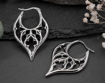 May include: A pair of silver hoop earrings with a gothic-inspired design. The earrings are made of metal and have a delicate, intricate pattern.