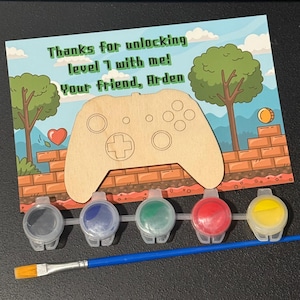 May include: A paint-your-own video game controller kit. The kit includes a wooden controller, paint pots in gray, blue, green, red, and yellow, and a paintbrush. The card reads, "Thanks for unlocking level 1 with me! Your friend, Arden."
