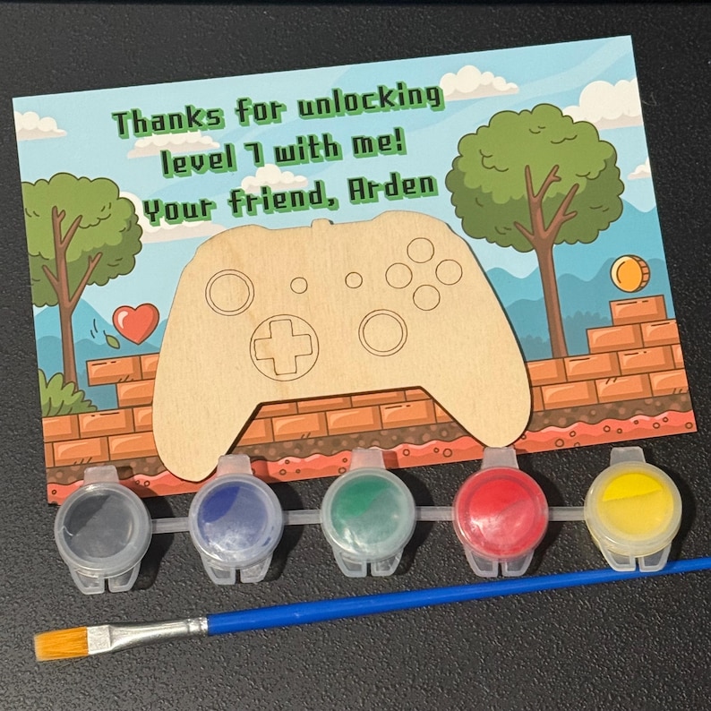 May include: A paint-your-own video game controller kit. The kit includes a wooden controller, paint pots in gray, blue, green, red, and yellow, and a paintbrush. The card reads, "Thanks for unlocking level 1 with me! Your friend, Arden."