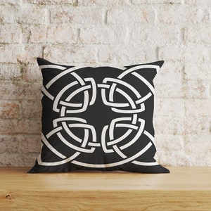 Peut inclure: Coussin carré noir avec un motif de nœud celtique blanc. Le coussin est posé sur une surface en bois devant un mur de briques blanches. Le motif géométrique est symétrique.