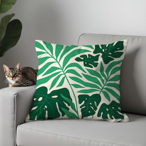 May include: A square cushion featuring a cream backdrop and a striking green leaf design. The cushion is positioned on a grey sofa. A tabby cat is resting on the sofa arm.