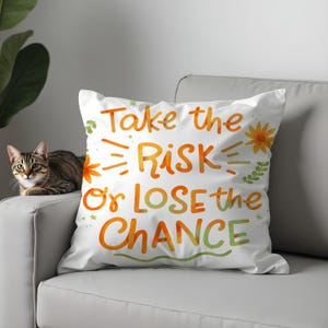 Può includere: Cuscino quadrato bianco con la scritta "Take the RISK or LOSE the CHANCE" in lettering acquerello arancione e verde. Il cuscino ha accenti floreali ed è su un divano grigio. Un gatto tigrato è sul divano.
