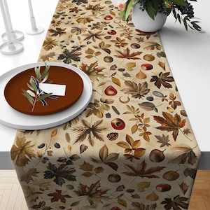 Puede incluir: Un camino de mesa beige con un diseño de otoño con hojas marrones, rojas y verdes, bayas y bellotas.