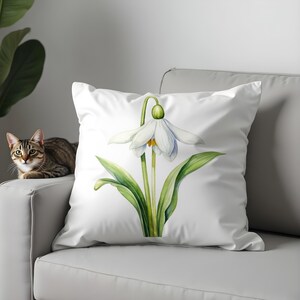 Può includere: Cuscino quadrato bianco con un'illustrazione ad acquerello di un fiore di bucaneve. Il fiore ha petali bianchi, un centro giallo e foglie e stelo verdi. Il cuscino è su un divano bianco, con un gatto sullo sfondo.