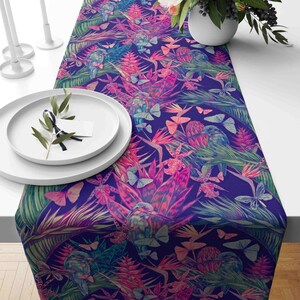 May include: A purple table runner with a tropical print featuring pink and green flowers, butterflies, and birds. The runner is made of fabric and has a soft, textured feel.