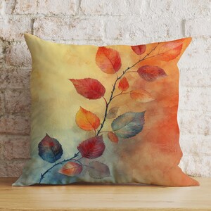 Könnte beinhalten: Ein dekoratives Kissen mit einem Aquarell-Design aus Herbstblättern in Rot-, Orange-, Gelb- und Blautönen auf einem Zweig. Der Hintergrund ist ein Farbverlauf von Gelb, Orange und Rot.