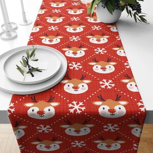 May include: Red table runner with a repeating pattern of white snowflakes and brown reindeer faces. The reindeer have white eyes, a red nose, and a white smile.