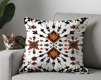 May include: White decorative throw pillow with a geometric pattern in orange, black, and brown. The pillow features a symmetrical design with diamond and arrow shapes. The pillow is on a white couch.