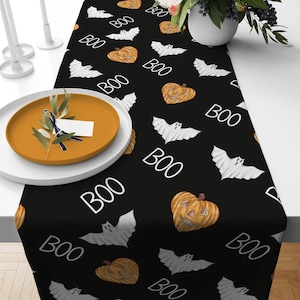 Może przedstawiać: Czarny bieżnik na stół na Halloween z powtarzającym się wzorem białych nietoperzy, pomarańczowych dyni i słowem "BOO" w kolorze białym. Idealny na imprezę Halloween, wykonany z miękkiej tkaniny.