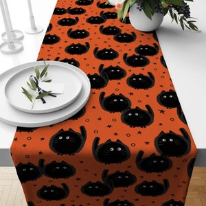 Puede incluir: Un camino de mesa naranja con un patrón de gatos negros. Los gatos son de estilo de dibujos animados y tienen ojos grandes. El fondo es naranja con estrellas y círculos blancos.