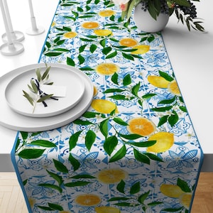 May include: A white table runner with a blue and white tile pattern and a lemon tree design. The lemons are yellow and the leaves are green.